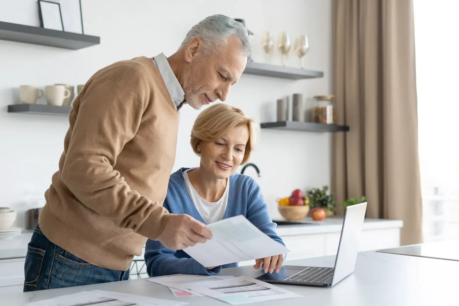 Smiling older couple reviewing financial documents together at home with a laptop, representing planning and understanding different types of insurance coverage.