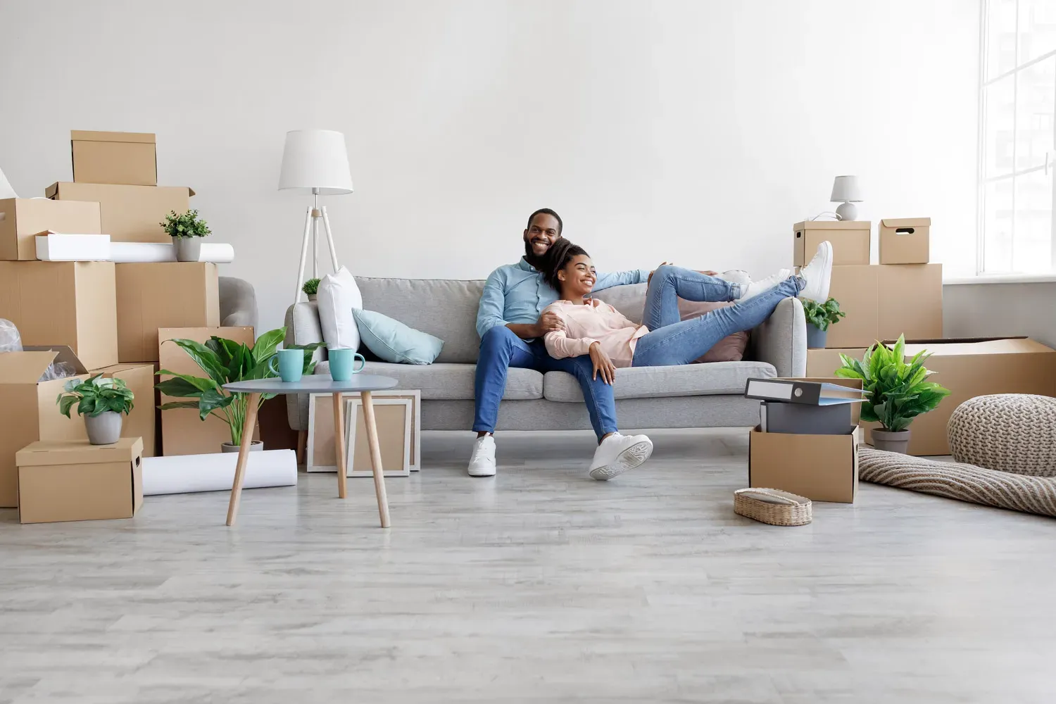 Smiling couple relaxing on a couch surrounded by moving boxes and plants, representing the excitement and success of first-time homebuyers settling into their new home.