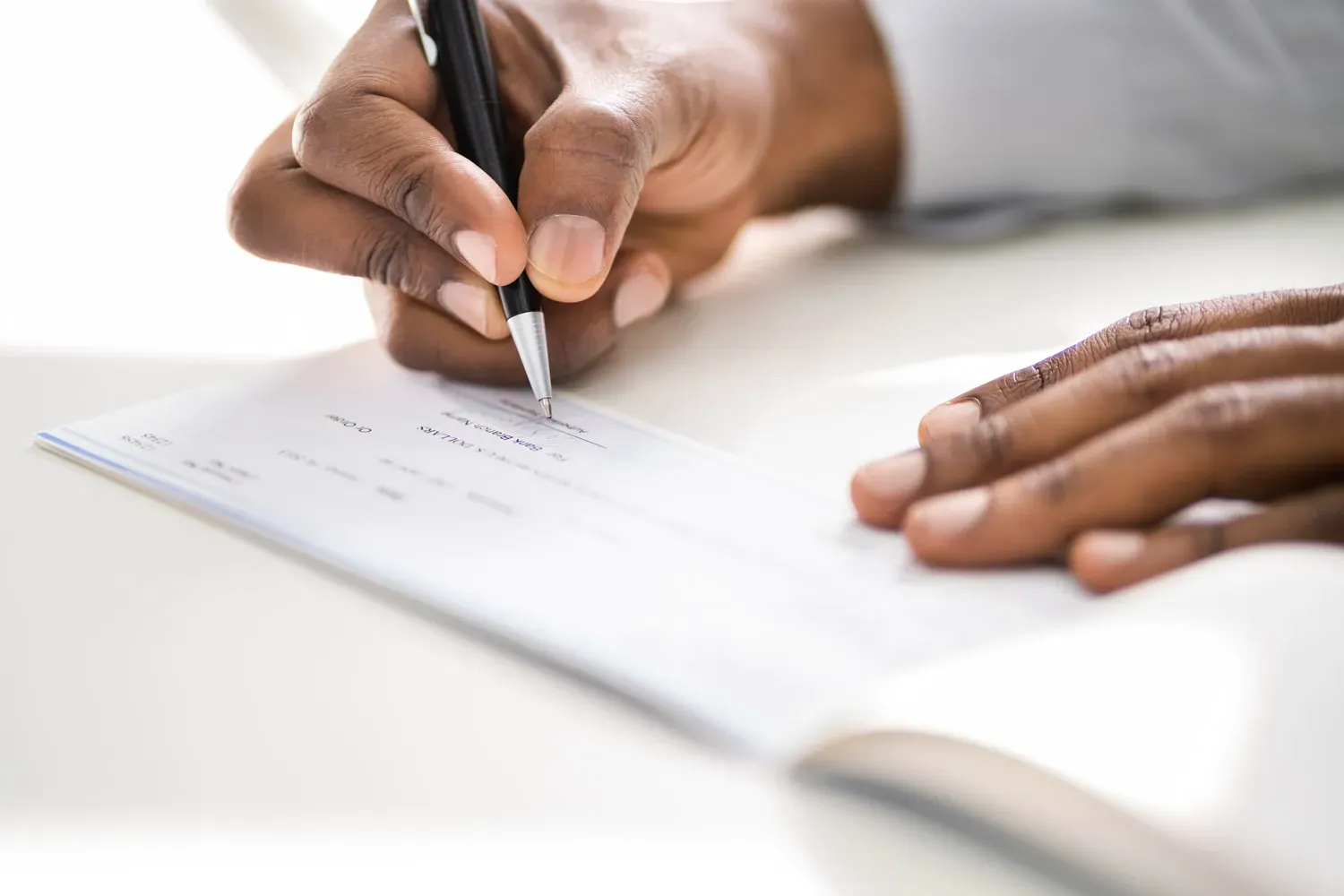 Close-up of a person's hands as they write on a check in a checkbook, with a pen poised over the signature line — illustrating cashing or filling out a check without using a bank.