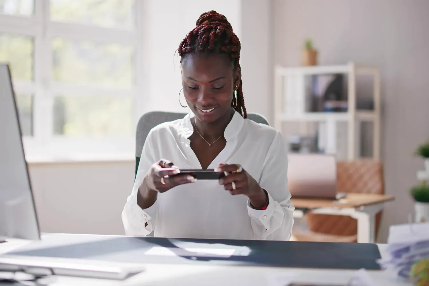Smiling woman sitting at her desk using her smartphone to deposit a check, representing the convenience and benefits of modern checking accounts.