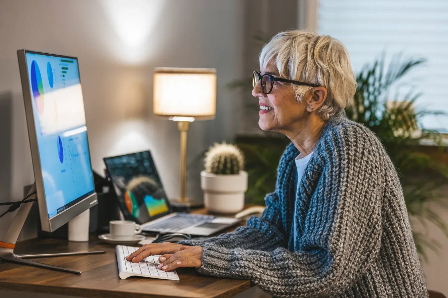 Smiling older woman reviewing financial charts on a computer, analyzing alternative investment options from home.