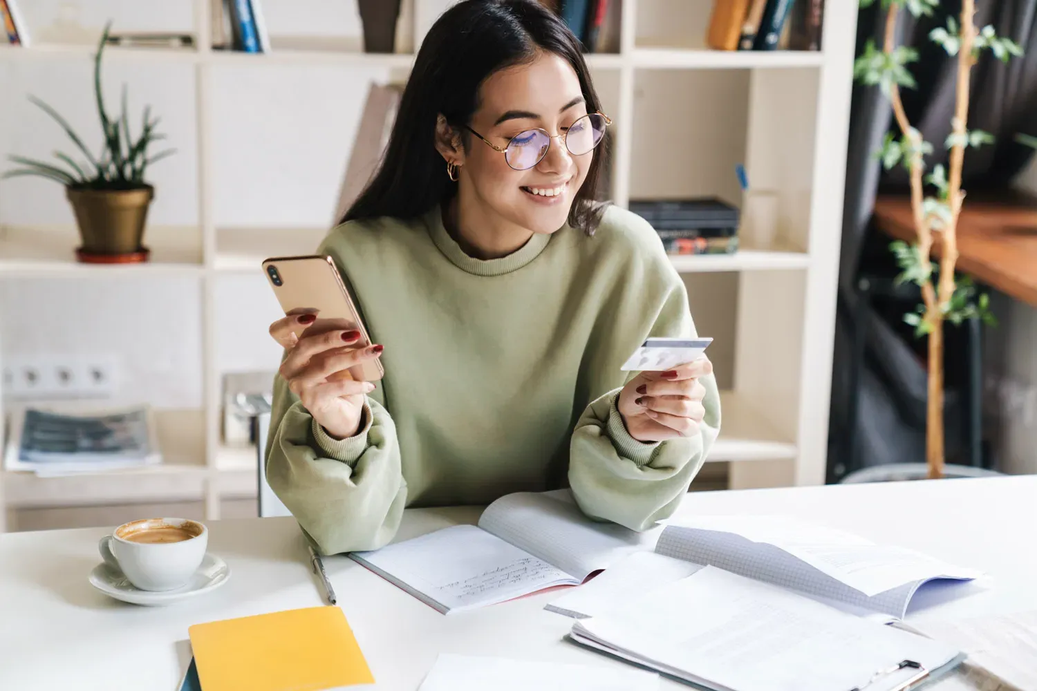 Smiling woman sitting at a desk holding her phone and credit card while reviewing tips to increase her chances of credit card application approval.