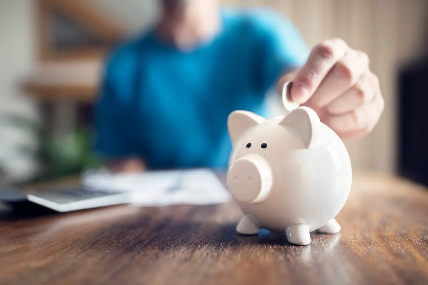 Person placing a coin into a white piggy bank on a wooden table, representing the benefits of having a savings account.