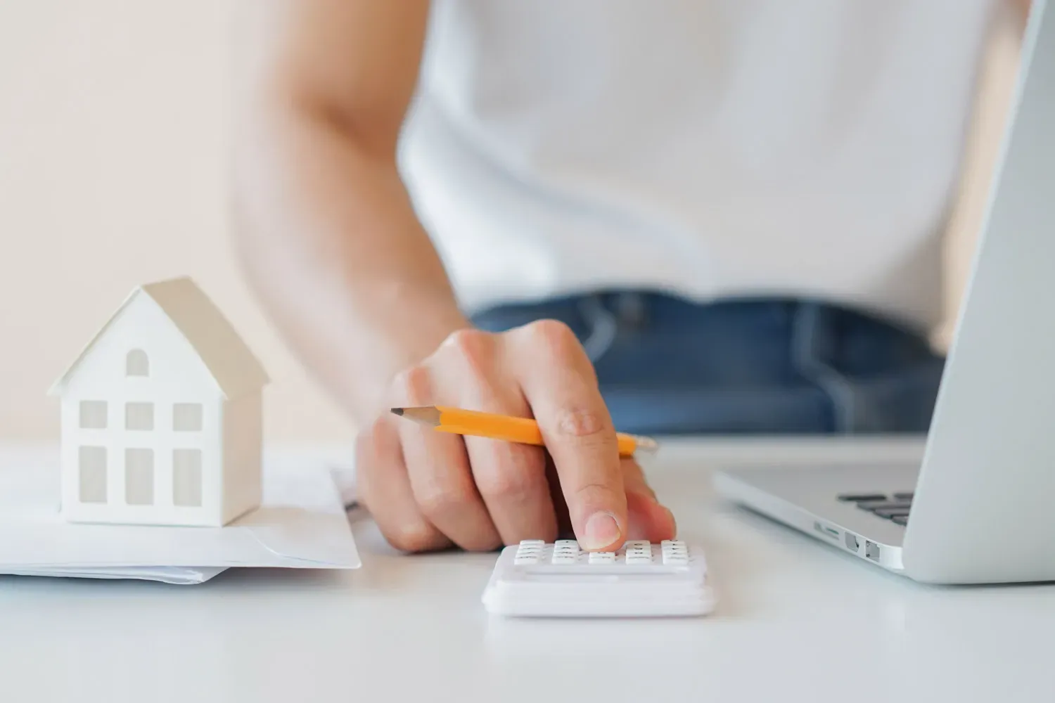 Person using a calculator with a small model house and laptop on the table, representing budgeting and financial planning for homeownership.