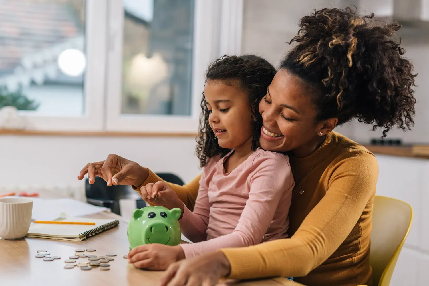 Smiling mother helping her young daughter put coins into a green piggy bank, representing the importance of teaching kids about savings through children’s savings accounts.