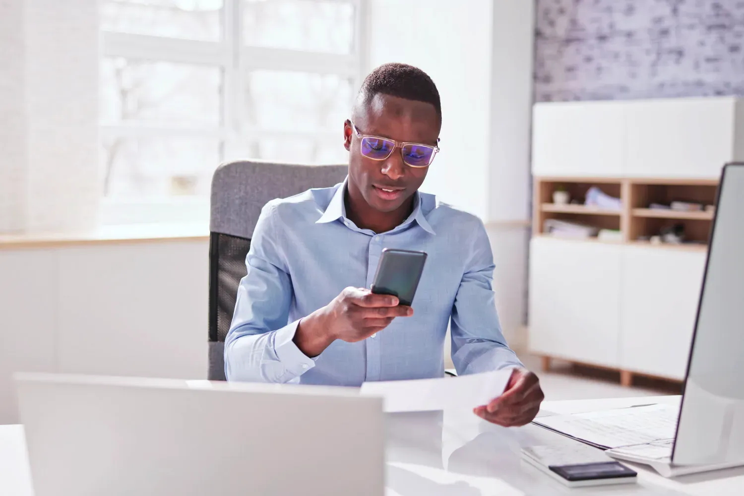 Man sitting at a desk using his smartphone to take a photo of a check for mobile deposit, representing modern checking account features like online and mobile banking.