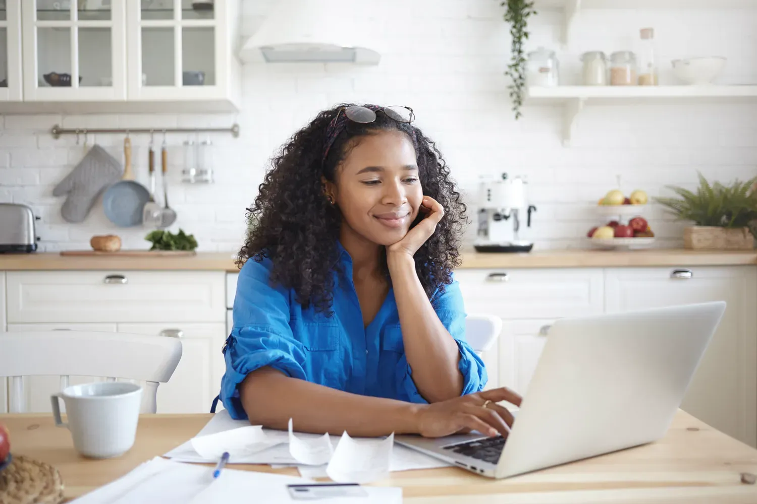 Woman sitting at a kitchen table using a laptop, reviewing bills and receipts with a calm expression — representing someone learning about overdraft protection and managing finances responsibly.