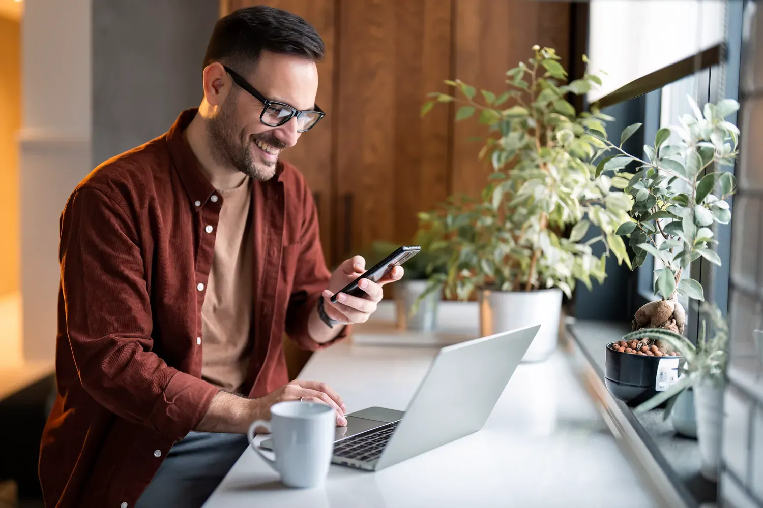Smiling man using his smartphone and laptop at a bright workspace, representing the convenience of managing different types of savings accounts online.