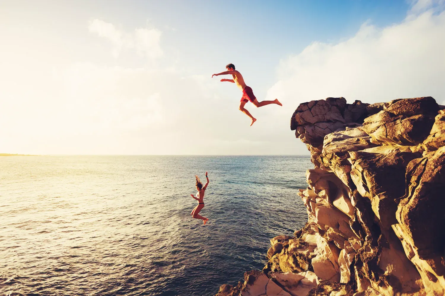 Two travelers cliff jumping into the ocean at sunset, symbolizing adventure and the importance of having travel insurance for unexpected events.