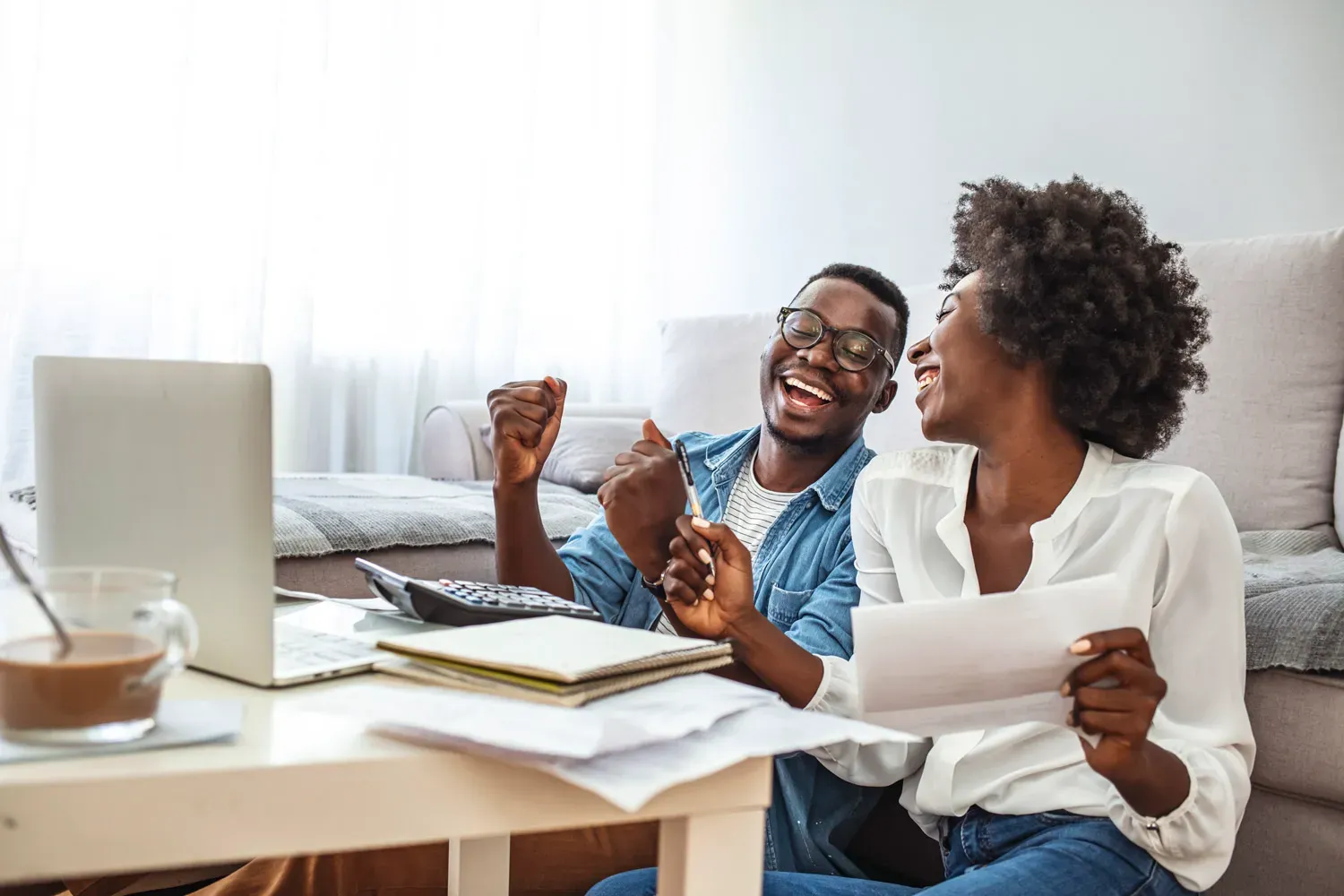 Happy couple celebrating while reviewing finances at home with a laptop and papers, symbolizing the benefits of choosing a high-quality savings account.