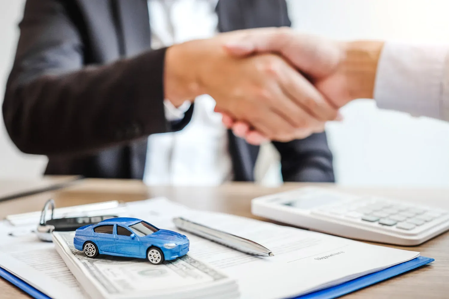 Close-up of two people shaking hands over car loan paperwork with a small blue toy car and money on the table, representing how to get approved for a car loan.