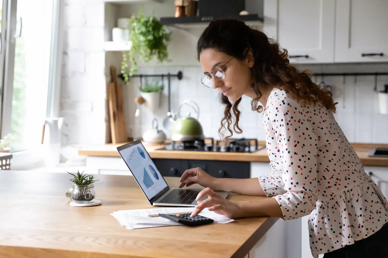 Young woman using a calculator and laptop to review charts and graphs, analyzing how the stock market works and tracking investments.