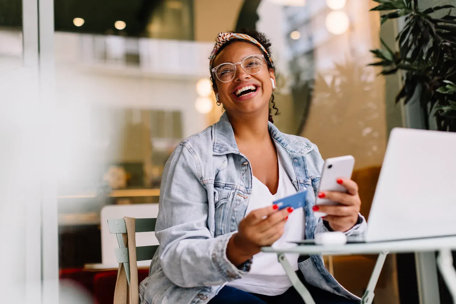 Woman sitting at an outdoor café smiling while holding a credit card and smartphone, with a laptop open in front of her as she applies for a new credit card online.