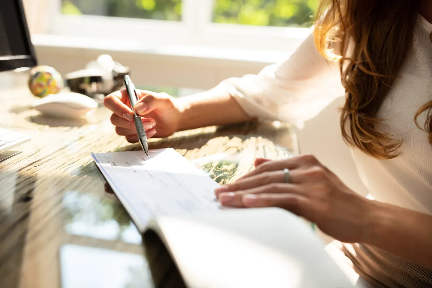 Close-up of a woman’s hands as she writes on a check at a sunlit desk, symbolizing the process of managing or closing a checking account.