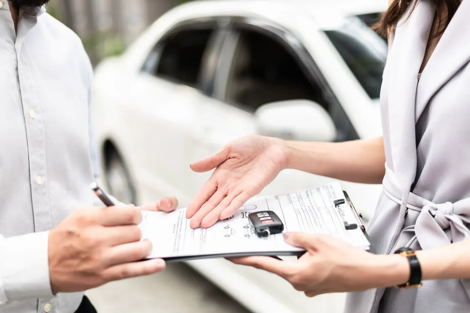 Person signing paperwork while another holds car keys and a contract, representing the process of refinancing a car loan to secure better terms.