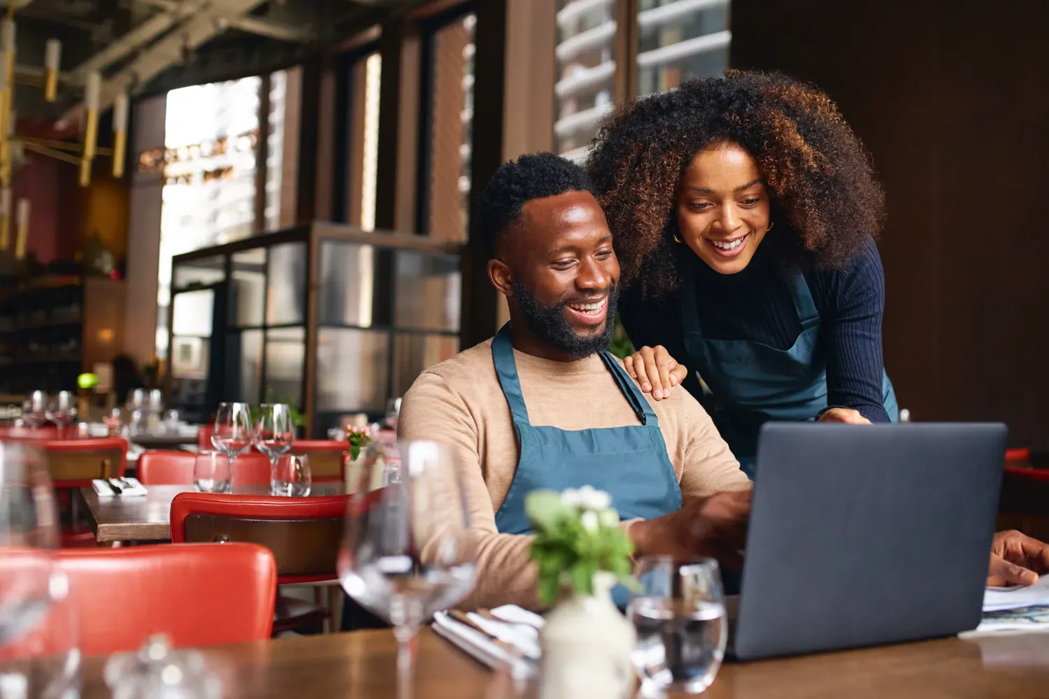 A smiling man and woman wearing aprons look at a laptop together inside a restaurant, suggesting small business owners setting up direct deposit for payroll or payments.