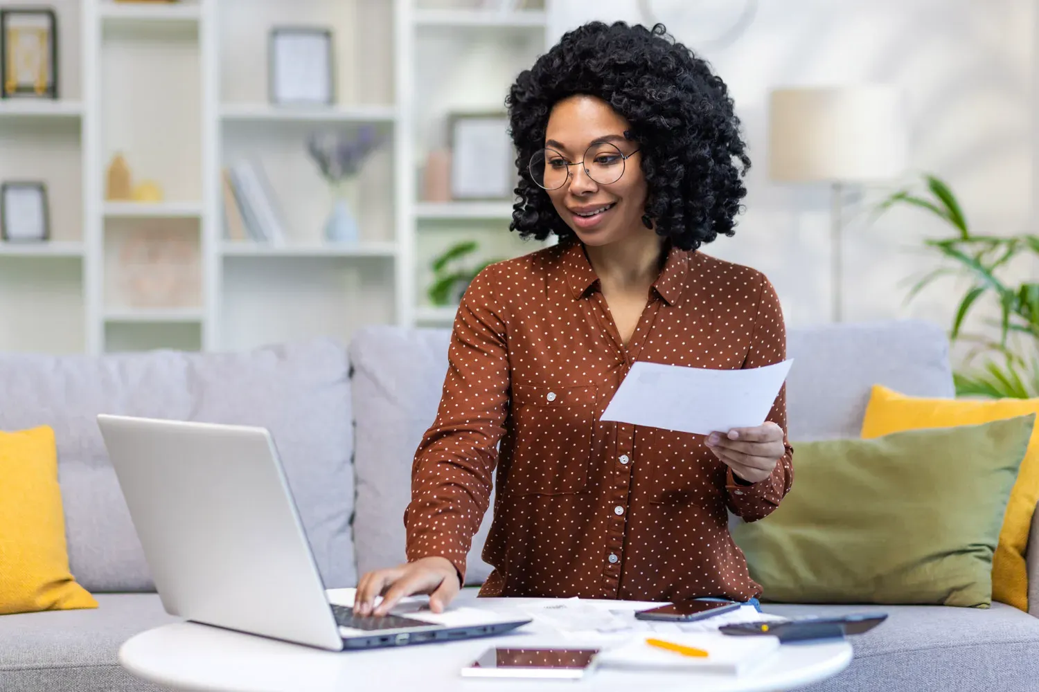 Smiling woman sitting on her couch while using a laptop and holding paperwork, representing someone managing finances and switching checking accounts with ease.
