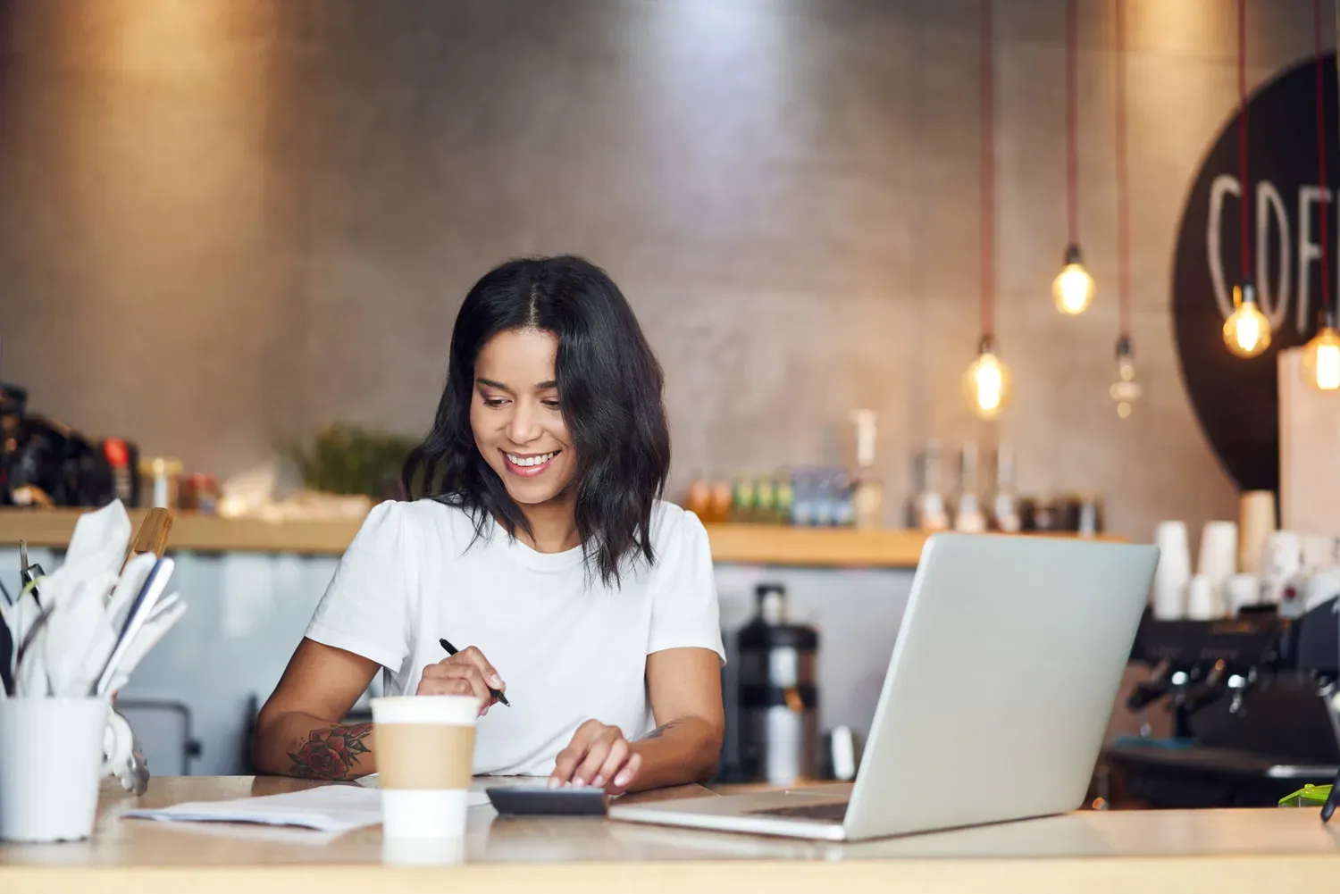 Smiling small business owner working on finances with a laptop and calculator in a café, representing a guide to using personal loans for small business growth.