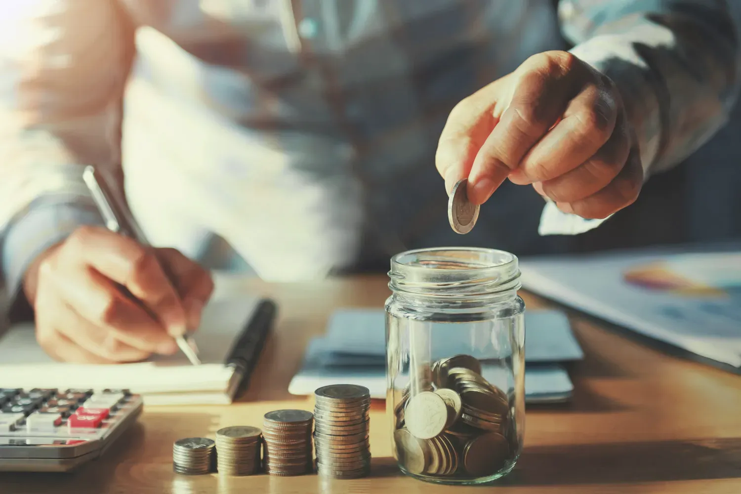 Person saving coins into a glass jar beside stacked coins, a notebook, and a calculator, symbolizing effective tips for growing personal savings.