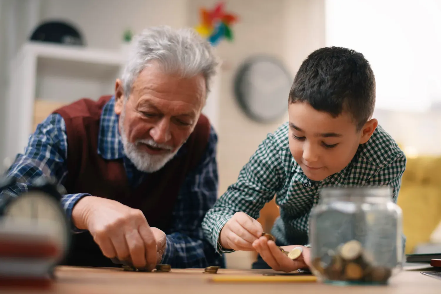 An elderly man and a young boy sitting together at a table, counting and stacking coins with a jar of savings nearby — symbolizing teaching children about money and financial responsibility.