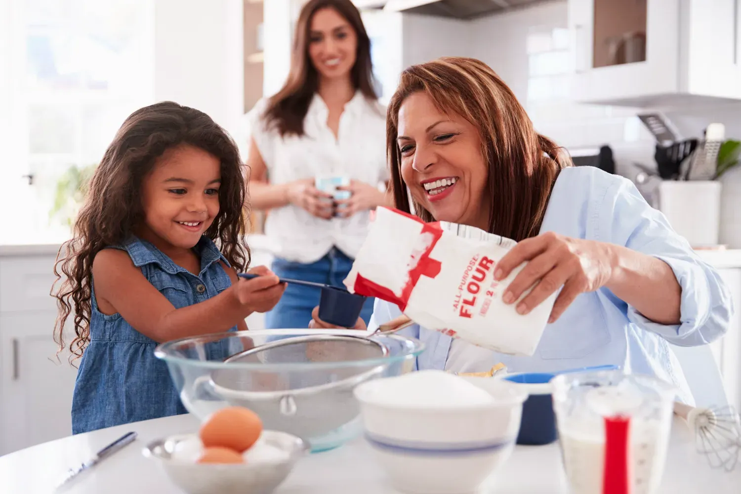 Grandmother baking with her granddaughter in the kitchen while a woman watches, representing family security and the long-term protection offered by life insurance.