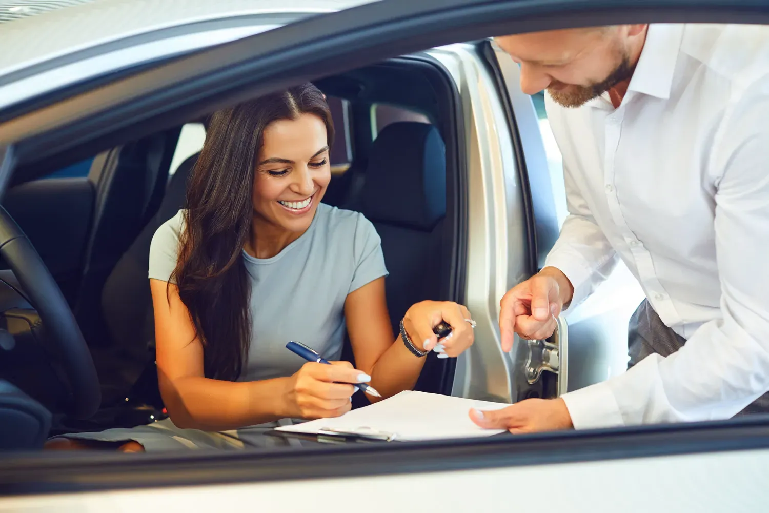 Smiling woman sitting in a car signing paperwork with a salesperson, representing calculating the true cost of owning and driving a vehicle.