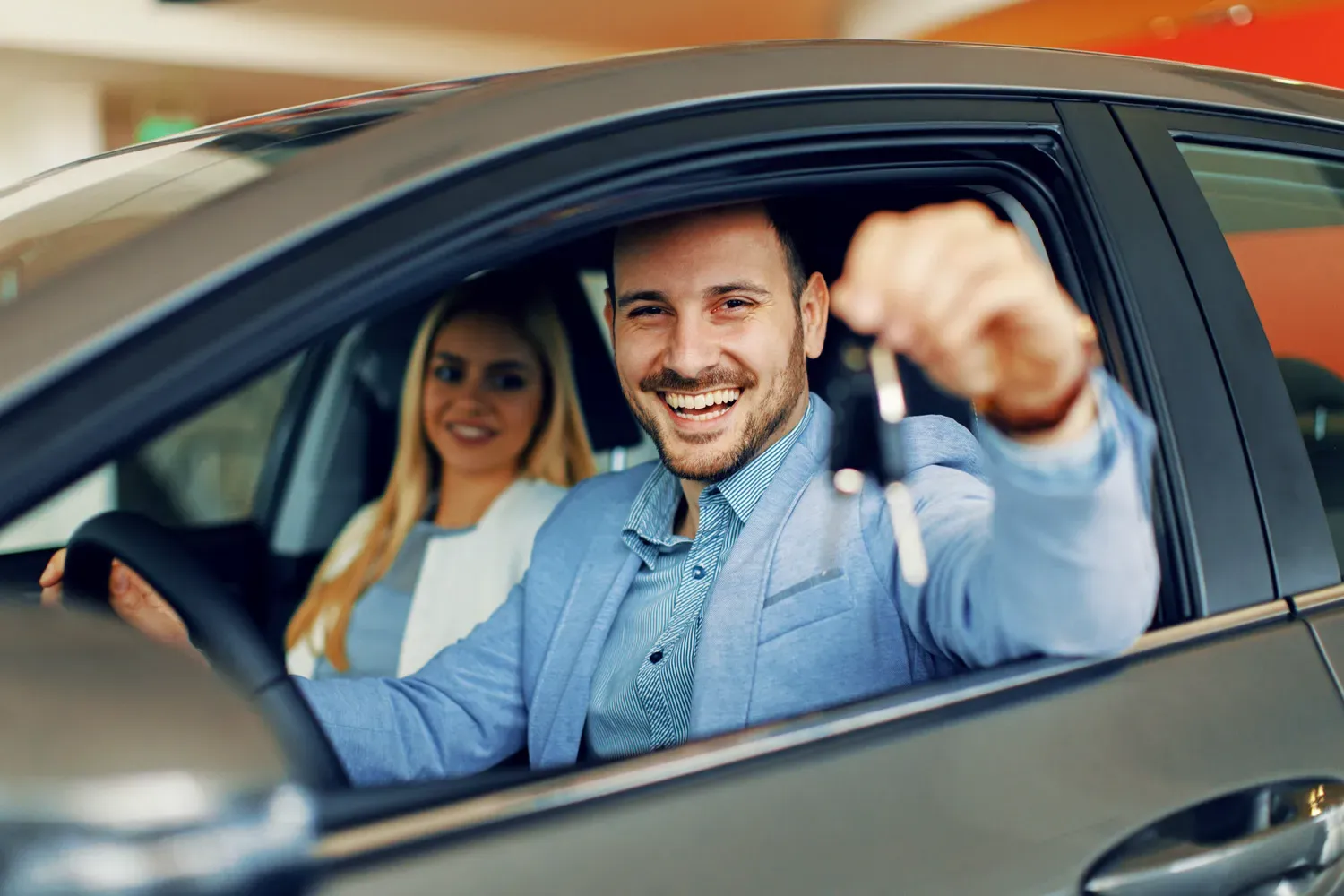 Smiling man sitting in a new car holding up car keys with a passenger beside him, representing alternatives to traditional car loans for vehicle financing.