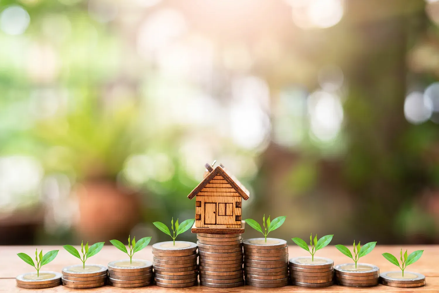 Stacks of coins with small plants growing on top and a miniature wooden house, symbolizing home equity growth and how property value builds financial wealth over time.