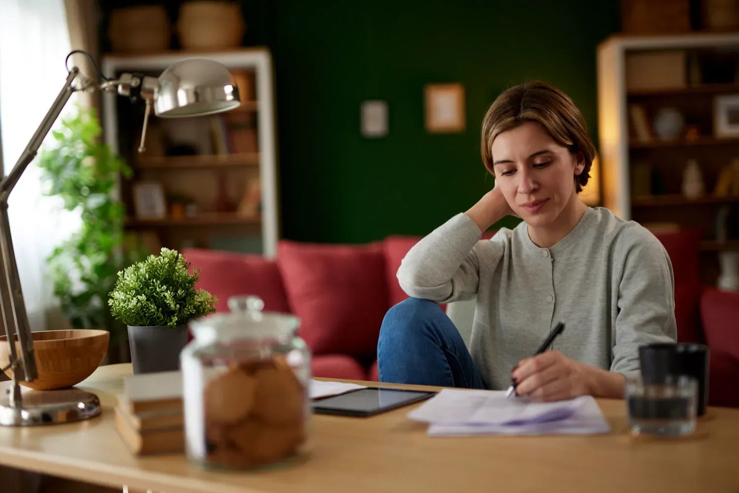 Woman sitting at a desk in her living room, reviewing papers and taking notes while deciding which type of checking account best fits her needs.